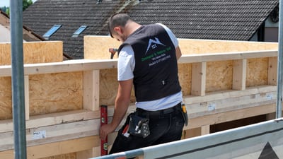 Ein Zimmerer der Zimmerei Wolf richtet auf dem Gerüst die Schwelle einer Holzrahmenwand mit der Wasserwaage aus; auf seiner Weste stehen Firmenlogo und „Wir leben Holz“.