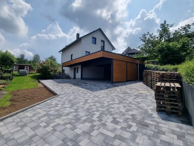 Wohnhaus mit angebautem Carport und großflächig gepflasterter Hoffläche, im Hintergrund der Hang mit Garten.
