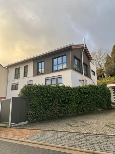 Dasselbe aufgestockte Wohnhaus von der Straße aus, hinter einer Efeuhecke, mit sichtbarem Holzdachüberstand.