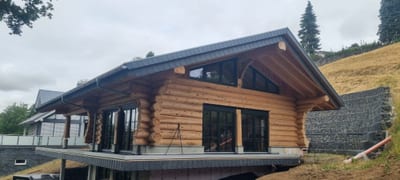 Fertiges Blockbohlenhaus mit weit auskragendem Satteldach, Schieferdeckung und geschwungenen Holzstützen an der Terrasse.