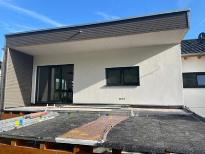 Anbau mit weit auskragendem Flachdach und dunkler Holz-Untersicht über einer Terrasse, daneben eine bodentiefe Schiebetür.