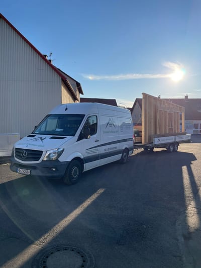 Transporter der Zimmerei Wolf mit Anhänger, auf dem ein vorgefertigtes Holzrahmen-Wandelement zur Baustelle gefahren wird.